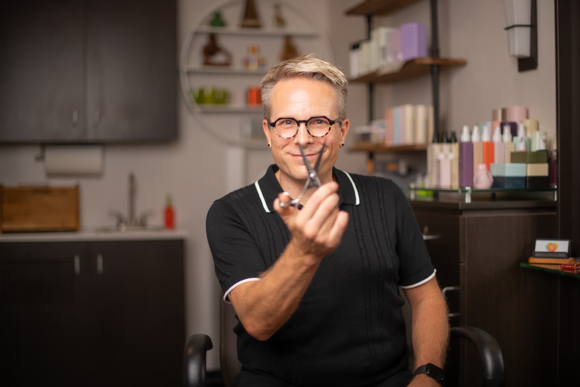 Ray Rose sitting in his Sunset Valley salon suite, smiling behind a pair of shears held up to the camera.